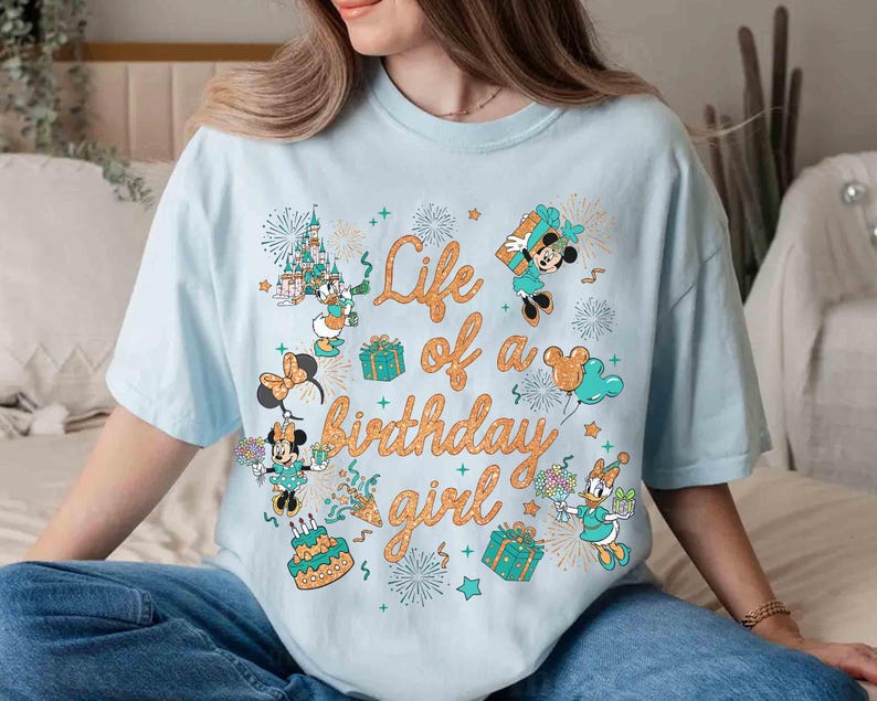 May include: Light blue t-shirt with the text "Life of a birthday girl" in orange script. The shirt features cartoon characters, including Mickey and Minnie Mouse, and Daisy Duck, along with birthday-themed graphics like fireworks, gifts, and a cake.