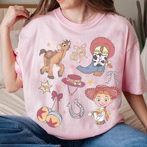 May include: Pale pink t-shirt with cartoon illustrations of a horse, cowboy boot, hat, and a cowgirl. The shirt has a crew neck and short sleeves. The design includes the word "Howdy" in a star-shaped frame.