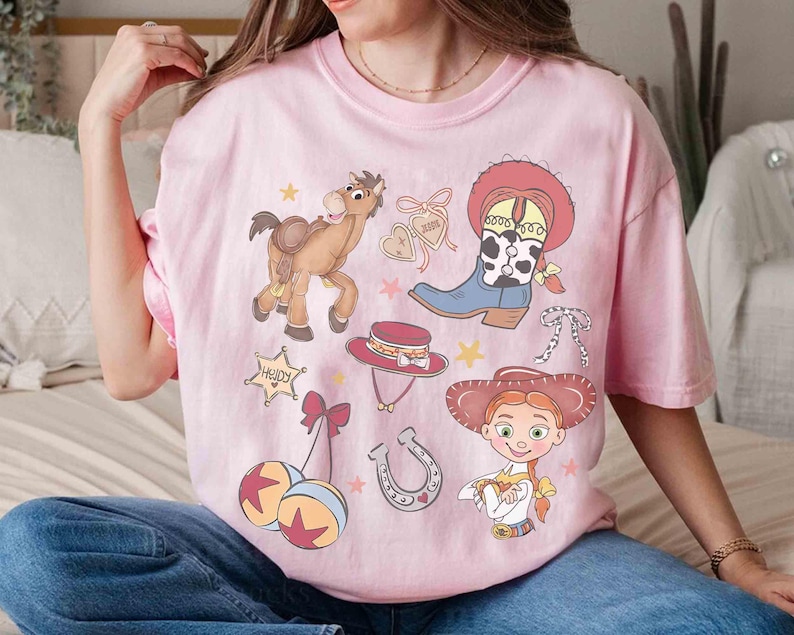 May include: Pale pink t-shirt with cartoon illustrations of a horse, cowboy boot, hat, and a cowgirl. The shirt has a crew neck and short sleeves. The design includes the word "Howdy" in a star-shaped frame.