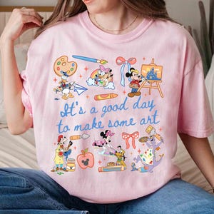 May include: Pink t-shirt with the text "It's a good day to make some art." Features cartoon characters like Mickey Mouse, Donald Duck, and others, engaged in art activities. Includes paint palettes, brushes, and a castle painting.