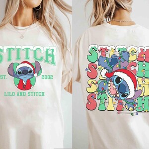 May include: Cream-colored t-shirt featuring a front graphic of Stitch wearing a Santa hat, with the text "STITCH" above and "LILO AND STITCH" below. The back of the shirt shows Stitch tangled in Christmas lights, with the word "STITCH" repeated.