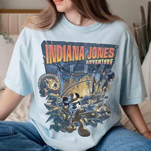 May include: A light blue t-shirt with a graphic print of Mickey Mouse dressed as Indiana Jones. The text "Indiana Jones Adventure" is printed above the image.