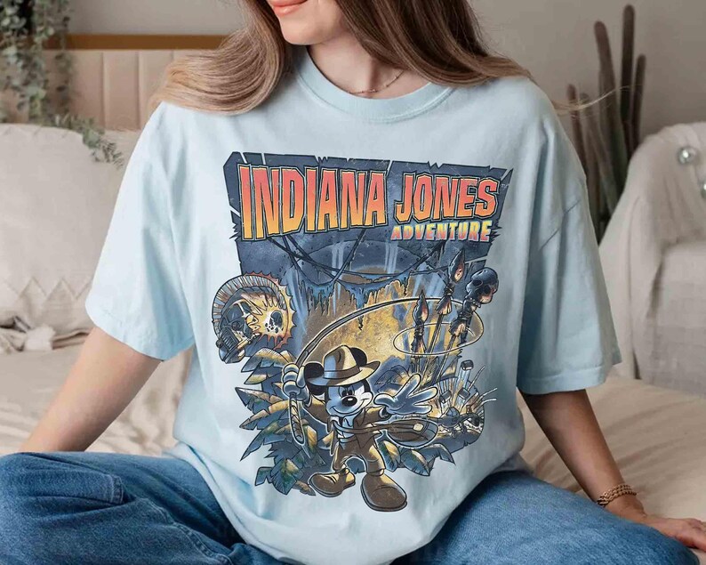 May include: A light blue t-shirt with a graphic print of Mickey Mouse dressed as Indiana Jones. The text "Indiana Jones Adventure" is printed above the image.
