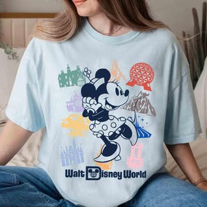 Może przedstawiać: Jasnoniebieska koszulka z grafiką Myszki Minnie w sukience w kropki, otoczonej kolorowymi ilustracjami zabytk&oacute;w Disney World. Tekst "Walt Disney World" jest wydrukowany poniżej na biało.