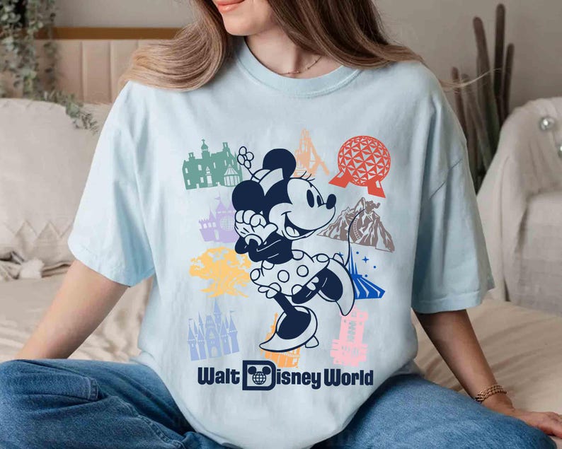 Może przedstawiać: Jasnoniebieska koszulka z grafiką Myszki Minnie w sukience w kropki, otoczonej kolorowymi ilustracjami zabytk&oacute;w Disney World. Tekst "Walt Disney World" jest wydrukowany poniżej na biało.