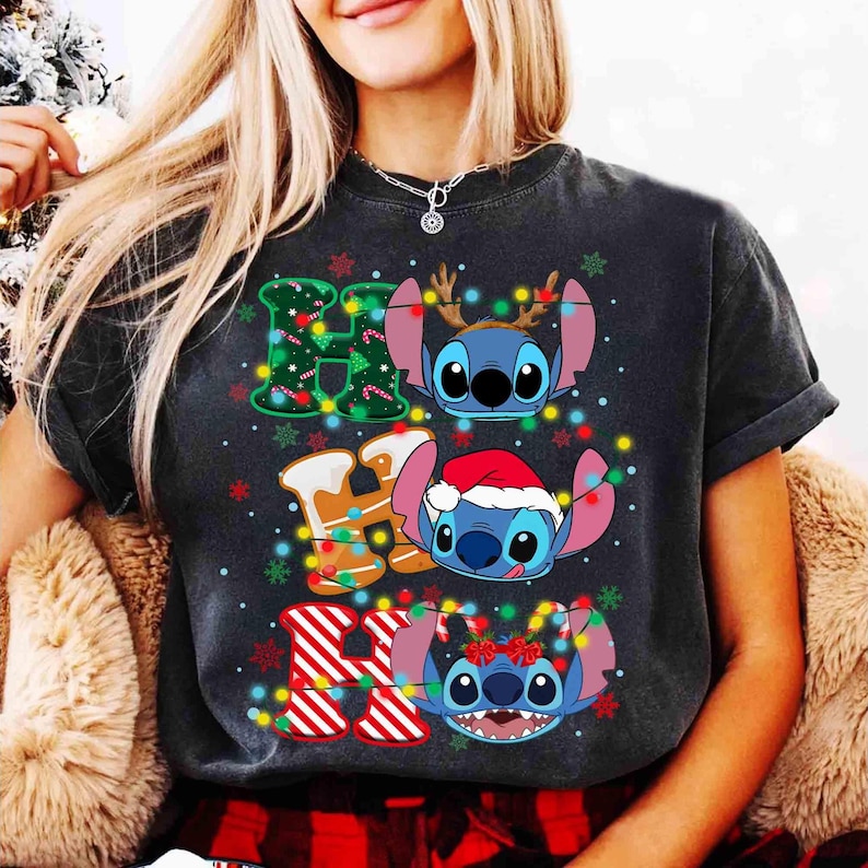 May include: Dark grey t-shirt with the word "HO" and Stitch characters. The "H" is decorated with Christmas lights and a Christmas tree design. The "O" is decorated with gingerbread and a Santa hat. The shirt has a festive holiday theme.