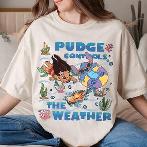 May include: Cream-colored t-shirt featuring a cartoon graphic of two characters swimming underwater with the text "Pudge Controls The Weather" in blue. The design includes cartoon fish, coral, and bubbles.