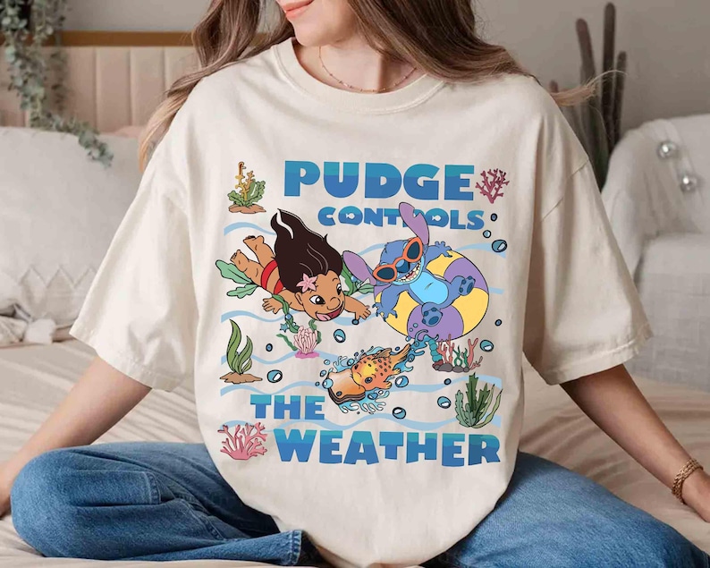 May include: Cream-colored t-shirt featuring a cartoon graphic of two characters swimming underwater with the text "Pudge Controls The Weather" in blue. The design includes cartoon fish, coral, and bubbles.