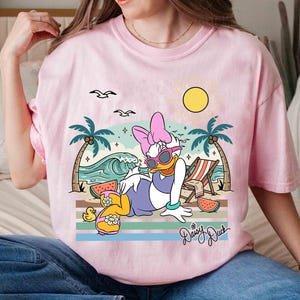 K&ouml;nnte beinhalten: Rosafarbenes T-Shirt mit einer Grafik von Daisy Duck, die sich am Strand entspannt, Sonnenbrille tr&auml;gt, mit Sonne, Palmen und Wellen. Der Schriftzug "Daisy Duck" ist kursiv. Das T-Shirt hat kurze &Auml;rmel und einen Rundhalsausschnitt.