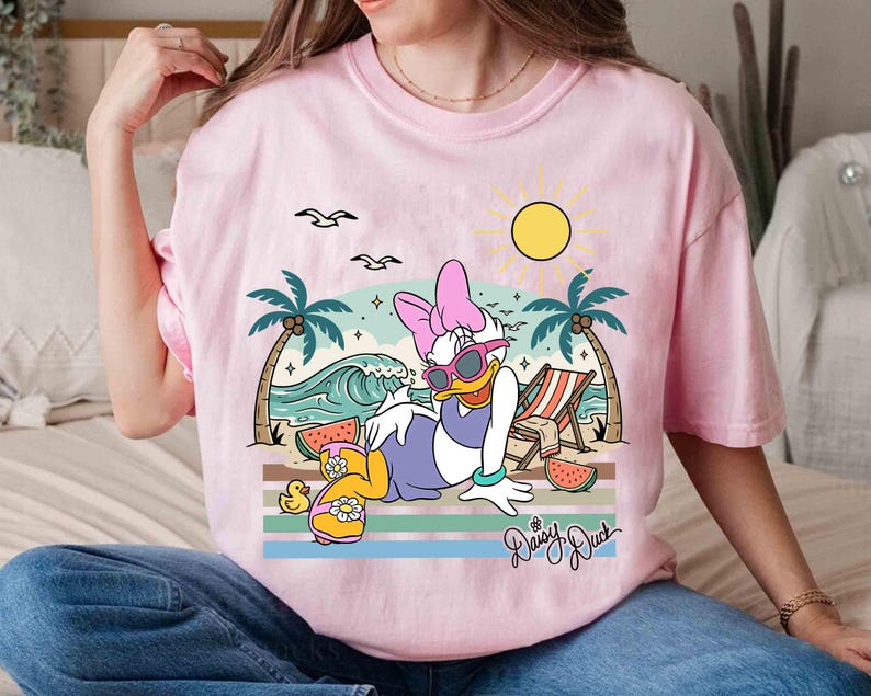 K&ouml;nnte beinhalten: Rosafarbenes T-Shirt mit einer Grafik von Daisy Duck, die sich am Strand entspannt, Sonnenbrille tr&auml;gt, mit Sonne, Palmen und Wellen. Der Schriftzug "Daisy Duck" ist kursiv. Das T-Shirt hat kurze &Auml;rmel und einen Rundhalsausschnitt.