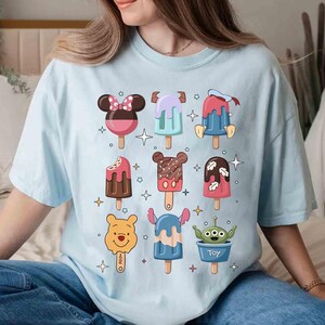 May include: Light blue t-shirt featuring colorful cartoon popsicle designs. The popsicles have various character-inspired designs, including Mickey Mouse, Stitch, and an alien. The shirt is a relaxed fit and suitable for casual wear.