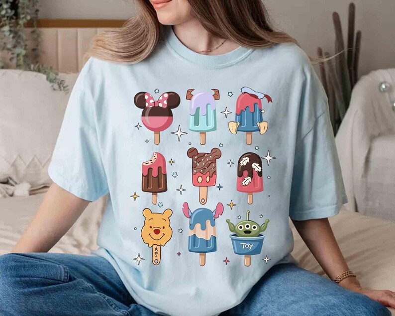 May include: Light blue t-shirt featuring colorful cartoon popsicle designs. The popsicles have various character-inspired designs, including Mickey Mouse, Stitch, and an alien. The shirt is a relaxed fit and suitable for casual wear.