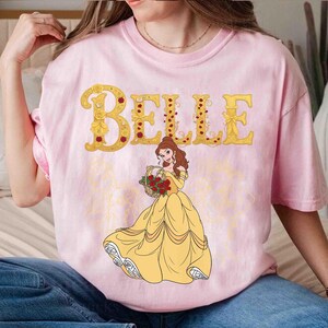 Op de afbeelding: Een lichtroze T-shirt met de naam "BELLE" in gouden letters en rode rozenaccenten. Het ontwerp bevat een cartoonillustratie van een vrouw in een gele jurk die een mand met rode rozen vasthoudt. Het T-shirt heeft een ronde hals.