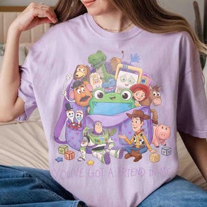 May include: A light purple t-shirt featuring a graphic print of various animated toy characters emerging from a purple backpack. The design includes a green frog, a space ranger, a cowboy, and other figures, with the text YOU'VE GOT A FRIEND IN ME.