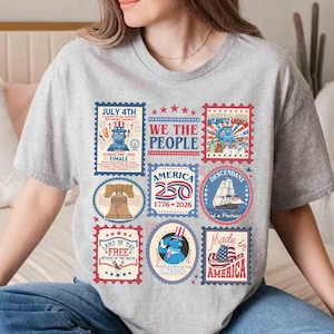 以下が含まれることがあります： 愛国的な切手デザインのコラージュが施されたグレーのTシャツ。切手には、自由の鐘、船、アンクル・サムの画像が含まれており、「July 4th」や「Made in America」などのテキストが記載されています。
