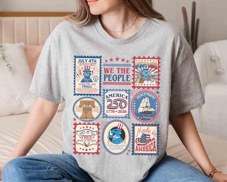 以下が含まれることがあります： 愛国的な切手デザインのコラージュが施されたグレーのTシャツ。切手には、自由の鐘、船、アンクル・サムの画像が含まれており、「July 4th」や「Made in America」などのテキストが記載されています。
