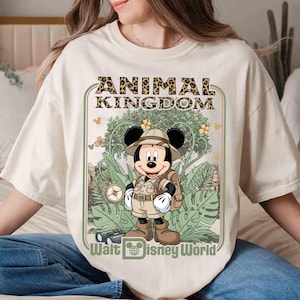 Puede incluir: Camiseta color crema con un dise&ntilde;o de Mickey Mouse con ropa de safari, el texto "ANIMAL KINGDOM" en estampado de leopardo y "Walt Disney World" debajo. El dise&ntilde;o incluye follaje de la jungla y una br&uacute;jula.