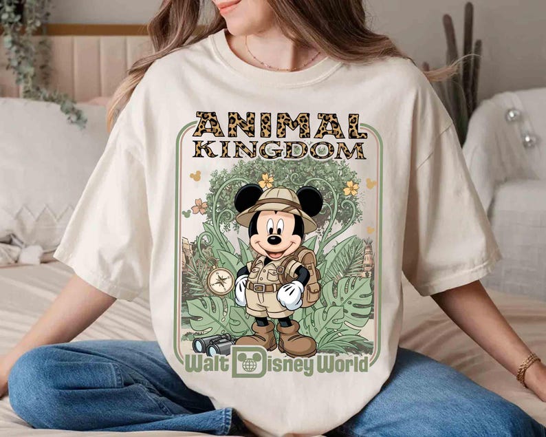 Puede incluir: Camiseta color crema con un dise&ntilde;o de Mickey Mouse con ropa de safari, el texto "ANIMAL KINGDOM" en estampado de leopardo y "Walt Disney World" debajo. El dise&ntilde;o incluye follaje de la jungla y una br&uacute;jula.