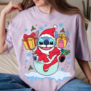 May include: A light purple t-shirt featuring a cartoon Stitch character dressed as Santa Claus, holding gifts. The design includes snowflakes and holly accents, creating a festive holiday theme.