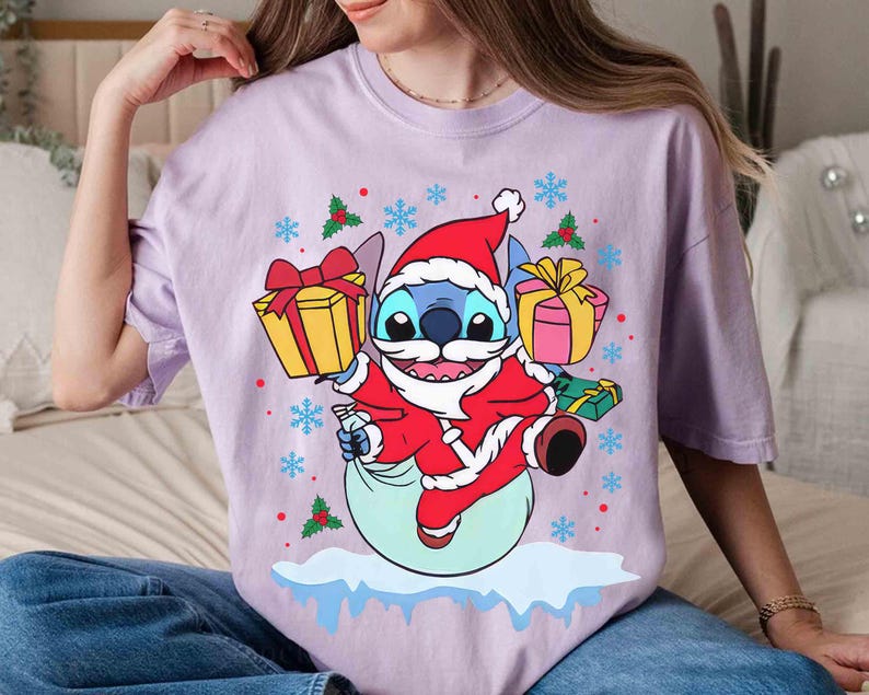 May include: A light purple t-shirt featuring a cartoon Stitch character dressed as Santa Claus, holding gifts. The design includes snowflakes and holly accents, creating a festive holiday theme.