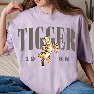 May include: A light purple t-shirt featuring the word "TIGGER" in gray block letters above a cartoon image of Tigger from Winnie the Pooh. The numbers "1968" are below the image. The shirt is a relaxed fit.
