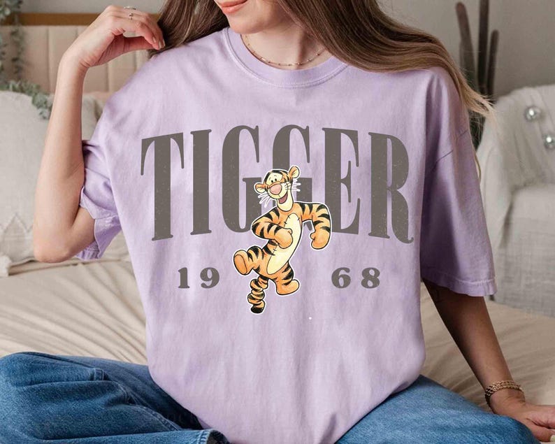 May include: A light purple t-shirt featuring the word "TIGGER" in gray block letters above a cartoon image of Tigger from Winnie the Pooh. The numbers "1968" are below the image. The shirt is a relaxed fit.