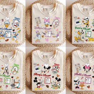 May include: Set of six cream-colored t-shirts featuring different Disney characters in a kitchen setting. Each shirt has a different character, including Mickey Mouse, Minnie Mouse, Donald Duck, Daisy Duck, Pluto, and Goofy. The shirts have a playful and colorful design with the text "[Character's name]'s Kitchen" on each shirt.