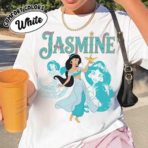 Op de afbeelding: Wit T-shirt met een afbeelding van prinses Jasmine die een gouden lamp vasthoudt, met de naam "Jasmine" in turquoise letters. Het shirt heeft ook turquoise illustraties van Jasmine. De woorden "Comfort Colors" en "White" staan in een cirkel.
