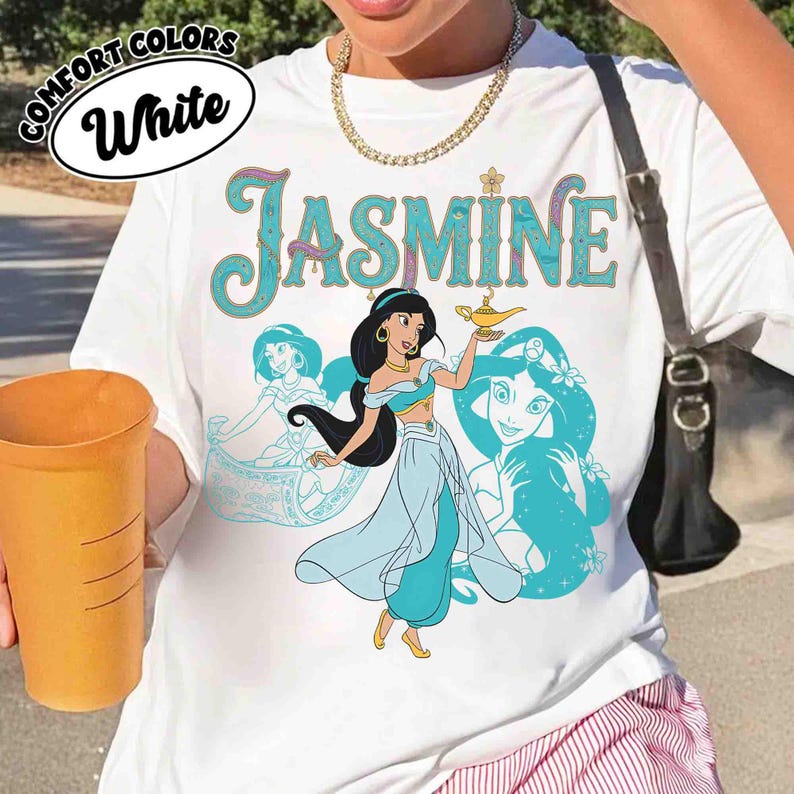 Op de afbeelding: Wit T-shirt met een afbeelding van prinses Jasmine die een gouden lamp vasthoudt, met de naam "Jasmine" in turquoise letters. Het shirt heeft ook turquoise illustraties van Jasmine. De woorden "Comfort Colors" en "White" staan in een cirkel.