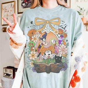 K&ouml;nnte beinhalten: Hellblaues T-Shirt mit einem farbenfrohen Aufdruck von Disney-Figuren, darunter Micky Maus, Minnie Maus, Donald Duck und Goofy, umgeben von Blumen, Flederm&auml;usen und einer schwarzen Katze. Das Design wird von einer gro&szlig;en Schleife und einem dekorativen Rahmen eingefasst.