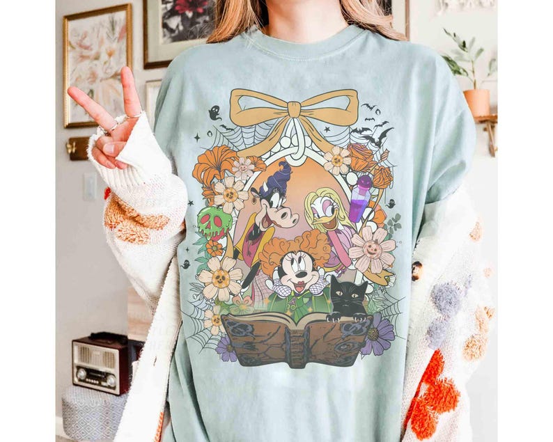 K&ouml;nnte beinhalten: Hellblaues T-Shirt mit einem farbenfrohen Aufdruck von Disney-Figuren, darunter Micky Maus, Minnie Maus, Donald Duck und Goofy, umgeben von Blumen, Flederm&auml;usen und einer schwarzen Katze. Das Design wird von einer gro&szlig;en Schleife und einem dekorativen Rahmen eingefasst.