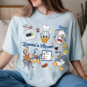 May include: A light blue t-shirt with a cartoon image of Donald Duck in a chef's hat. The shirt has the text "Donald's Kitchen" and features other cartoon images of Donald Duck cooking and grilling.