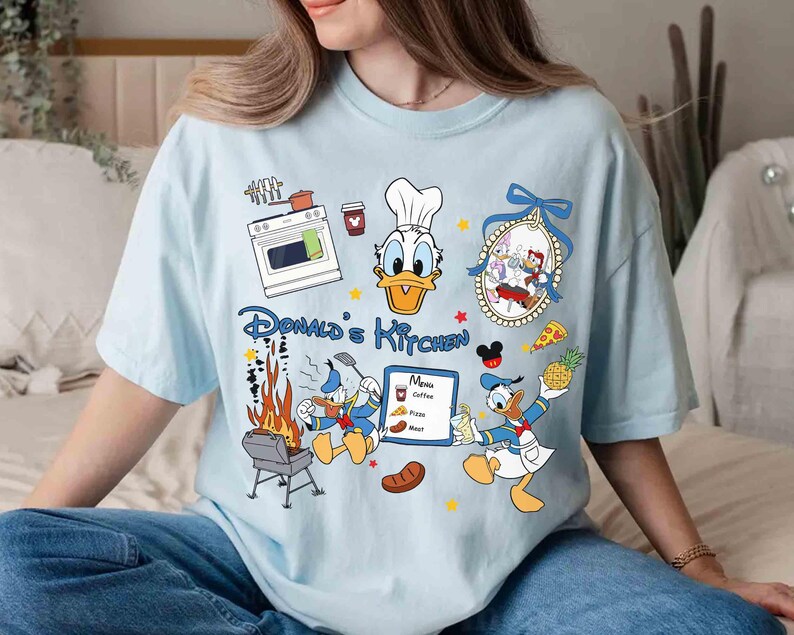 May include: A light blue t-shirt with a cartoon image of Donald Duck in a chef's hat. The shirt has the text "Donald's Kitchen" and features other cartoon images of Donald Duck cooking and grilling.