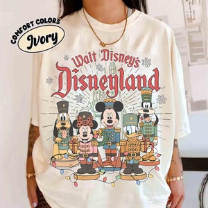 May include: Ivory-colored t-shirt featuring a graphic of Disney characters dressed as toy soldiers. The text "Walt Disney's Disneyland" is displayed in red and green. The shirt is a Comfort Colors brand.