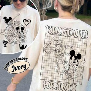 Puede incluir: Camiseta color marfil con gr&aacute;ficos en blanco y negro de Kingdom Hearts. La parte delantera presenta un dise&ntilde;o circular con personajes y s&iacute;mbolos. La parte trasera muestra el texto "KINGDOM HEARTS". La camiseta es de Comfort Colors.