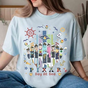 K&ouml;nnte beinhalten: Hellblaues T-Shirt mit einem farbenfrohen Grafikdesign von Pixar-Charakteren in Matrosenuniformen, mit dem Text "PIXAR Day At Sea". Das Design beinhaltet nautische Elemente wie ein Steuerrad, Anker und M&ouml;wen.