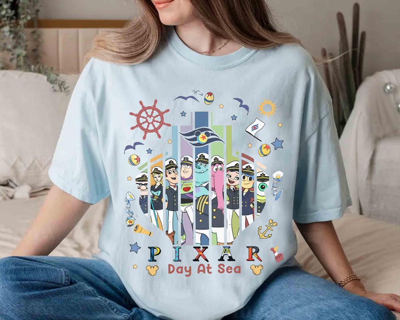 K&ouml;nnte beinhalten: Hellblaues T-Shirt mit einem farbenfrohen Grafikdesign von Pixar-Charakteren in Matrosenuniformen, mit dem Text "PIXAR Day At Sea". Das Design beinhaltet nautische Elemente wie ein Steuerrad, Anker und M&ouml;wen.