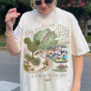 May include: Cream-colored t-shirt featuring a colorful graphic of Disney characters on a boat ride. The design includes the text "Only then will we truly" and "Living with the Land". The shirt has a relaxed fit.