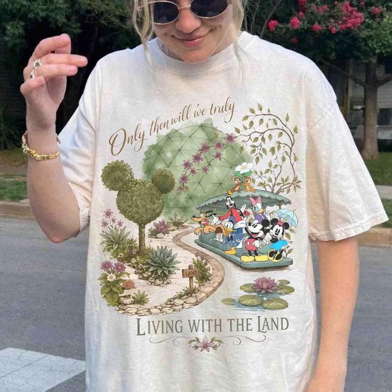 May include: Cream-colored t-shirt featuring a colorful graphic of Disney characters on a boat ride. The design includes the text "Only then will we truly" and "Living with the Land". The shirt has a relaxed fit.