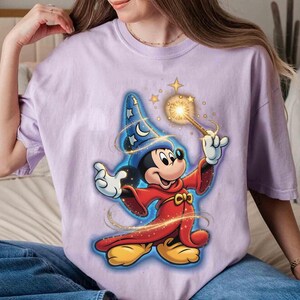 May include: A light purple t-shirt featuring a cartoon wizard Mickey Mouse design. Mickey is wearing a blue wizard hat with stars and a red robe, holding a glowing wand. The design has gold accents and stars.