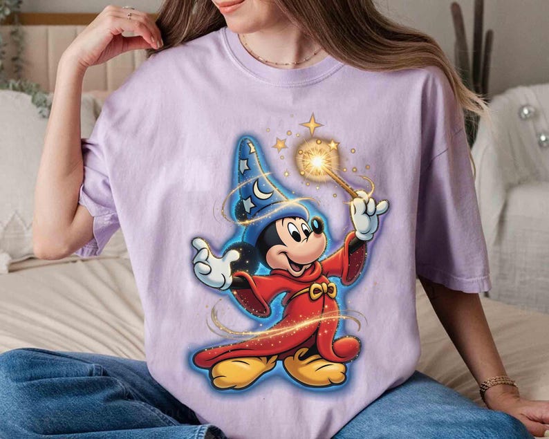 May include: A light purple t-shirt featuring a cartoon wizard Mickey Mouse design. Mickey is wearing a blue wizard hat with stars and a red robe, holding a glowing wand. The design has gold accents and stars.