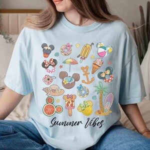 以下が含まれることがあります： 明るいブルーのTシャツに、さまざまなディズニーキャラクターと夏のテーマのアイコンがカラフルなグラフィックプリントされています。デザインの下部には「Summer Vibes」というテキストが黒で印刷されています。