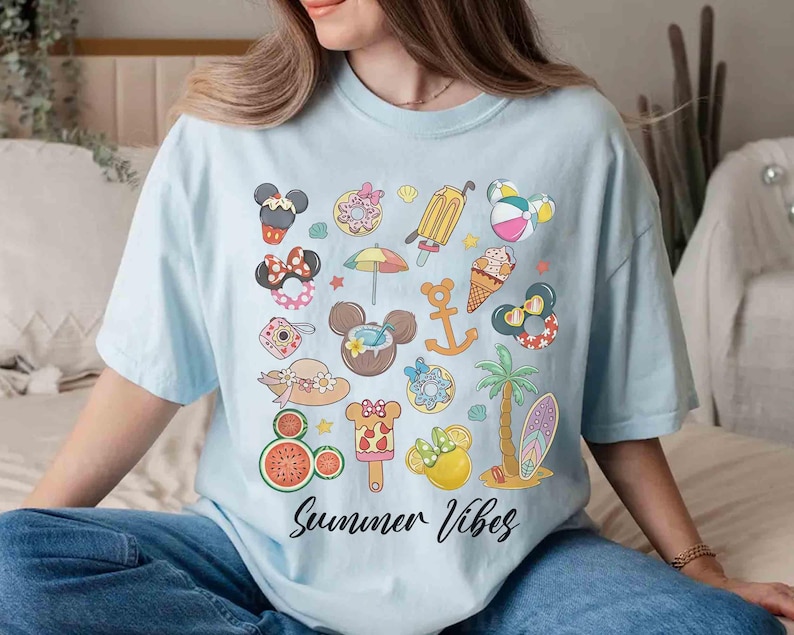 以下が含まれることがあります： 明るいブルーのTシャツに、さまざまなディズニーキャラクターと夏のテーマのアイコンがカラフルなグラフィックプリントされています。デザインの下部には「Summer Vibes」というテキストが黒で印刷されています。