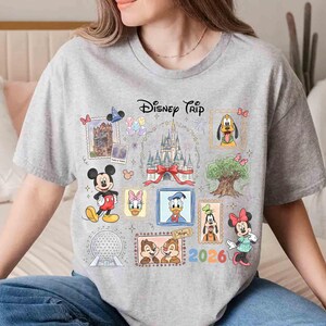 Puede incluir: Una camiseta gris con un dise&ntilde;o colorido de "Disney Trip". El dise&ntilde;o incluye varios personajes y lugares emblem&aacute;ticos de Disney, como Mickey Mouse, Minnie Mouse y el castillo de Cenicienta. Tambi&eacute;n aparece el a&ntilde;o 2026.