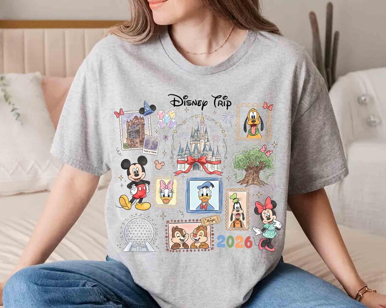 Puede incluir: Una camiseta gris con un dise&ntilde;o colorido de "Disney Trip". El dise&ntilde;o incluye varios personajes y lugares emblem&aacute;ticos de Disney, como Mickey Mouse, Minnie Mouse y el castillo de Cenicienta. Tambi&eacute;n aparece el a&ntilde;o 2026.