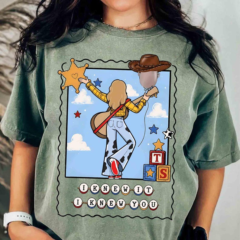 May include: A sage green t-shirt featuring a graphic of a person with blonde hair wearing a yellow plaid shirt and cow-print pants, holding a guitar and a brown cowboy hat balloon. The text "I KNEW IT I KNEW YOU" is printed below the illustration.