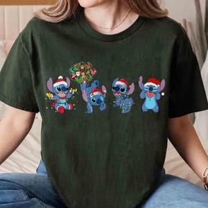 Puede incluir: Camiseta verde oscuro con un dise&ntilde;o navide&ntilde;o que presenta cuatro personajes de dibujos animados azules, cada uno con un gorro de Pap&aacute; Noel y luces festivas. Los personajes son de la pel&iacute;cula animada Lilo & Stitch. El dise&ntilde;o est&aacute; impreso en la parte delantera.