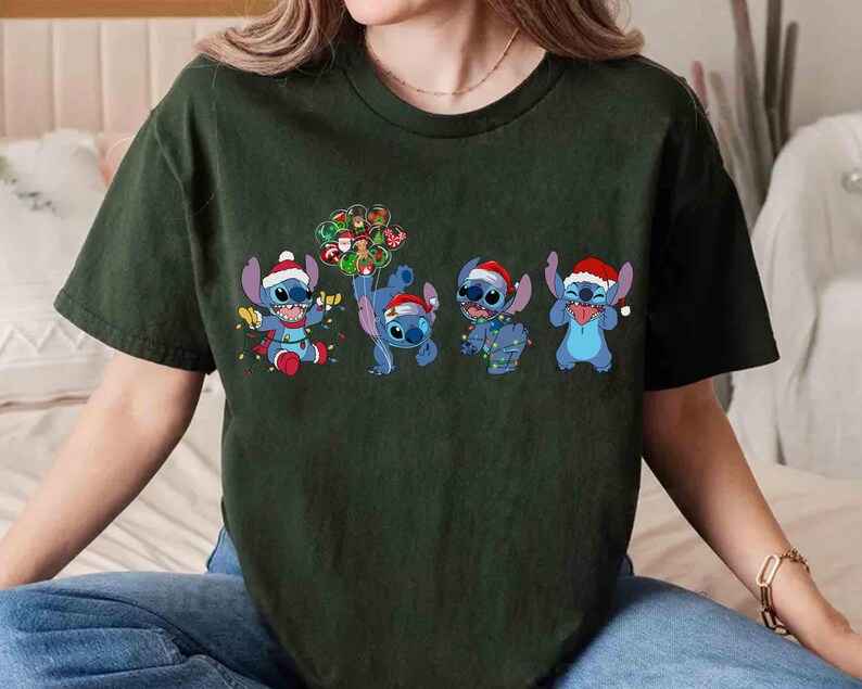 Puede incluir: Camiseta verde oscuro con un dise&ntilde;o navide&ntilde;o que presenta cuatro personajes de dibujos animados azules, cada uno con un gorro de Pap&aacute; Noel y luces festivas. Los personajes son de la pel&iacute;cula animada Lilo & Stitch. El dise&ntilde;o est&aacute; impreso en la parte delantera.