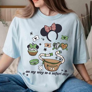 K&ouml;nnte beinhalten: Hellblaues T-Shirt mit einem Safari-Themen-Grafikdesign, das Minnie-Maus-Ohren, eine Bauchtasche und andere Elemente zeigt. Der Text "on my way to a safari day" ist auf dem Shirt aufgedruckt. Das Shirt hat eine lockere Passform.