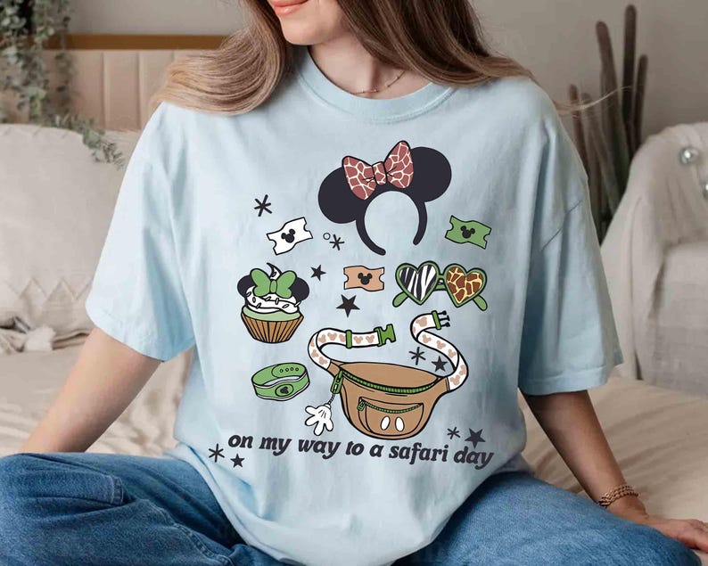 K&ouml;nnte beinhalten: Hellblaues T-Shirt mit einem Safari-Themen-Grafikdesign, das Minnie-Maus-Ohren, eine Bauchtasche und andere Elemente zeigt. Der Text "on my way to a safari day" ist auf dem Shirt aufgedruckt. Das Shirt hat eine lockere Passform.